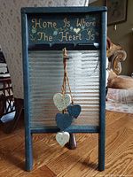 Front view of the wooden framed washboard showing the painted folk art lettering and floral decorations above the glass scrubbing surface with four hanging hearts suspended by rope.