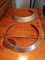 Top-down view showing both vintage brass chokers placed flat on wooden surface, highlighting one hammered and one ribbed metal finish.