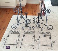 Six black metal plate holders on a light countertop, showing 3 larger easel stands with curled decorative metalwork and 3 smaller wall-mounted holders with hooks.
