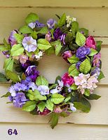 Full view of the circular floral wreath with a variety of purple, pink, and lavender flowers and bright green leaves on a beige siding background.