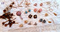 Overview of 19 pairs of clip-on earrings laid out on a white quilted surface with a ruler for scale. Earring styles range from floral clusters with colorful rhinestones to gold-tone metal designs and pearl clusters.