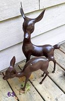 Photo of two dark brown spray painted metal deer figurines, one standing and one grazing on wooden surface against light siding.