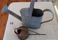 Photo of the larger galvanized metal watering can with a long spout and curved handle next to the smaller brass-colored watering can on a white quilted surface.