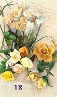 Lot of twelve yellow, cream, and beige porcelain roses with some mounted on green stems and leaves, arranged on a cloth background.