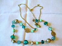 Full view of the necklace laid out on a white surface showing the array of teal, turquoise, and gold-tone beads with chain and clasp.