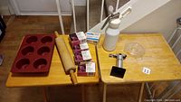 Photo showing a collection of baking items on two small tables, including silicone muffin molds, wooden rolling pin, soda chargers, soda siphon dispenser, metal creamer, creme brulee burner, and glass lemon juicer.