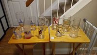 Group photo of 12 glass beer mugs and collectible glasses featuring Disney characters and other designs on two wooden tables.