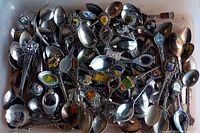 Multiple collectors spoons with crest and emblem decorations in a box, showing tarnish and scratches.