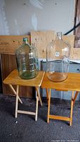 Two large glass bottles on small folding wooden tables, one clear and one greenish glass jug, used for wine making.