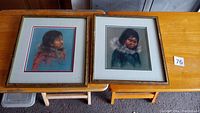 Two framed Inuit child portraits side-by-side, gold colored wooden frames, mats, on wood tables.