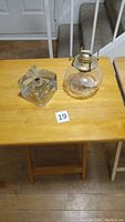 Two oil lamps on wooden table, one cube-shaped clear glass and one round clear glass with brass burner.