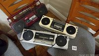 Three radio cassette players displayed on chairs; maroon Sanyo, silver Panasonic and off-white Panasonic