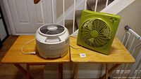 Photo shows white room humidifier and green box fan side by side on wooden tables, visible cords included.