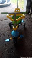 Front view of the SmarTrike convertible tricycle showing yellow seat with green handle, blue front wheel, pedals and footrest, green handle grips on silver metal handlebars.