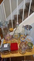 Top-down view of 13 assorted glass containers on a wooden surface near a stairway. Includes various jars and bottles, some with lids, swing-top stoppers and corks.
