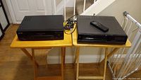 Two black VHS players on wooden tables, one Sharp and one GE with remote on top