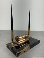 Photo showing side angle of two black fountain pens with gold-colored tips inserted in gold holders on black stone base with rotary calendar.