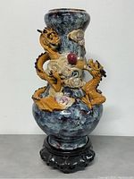 Full frontal view of the pottery vase with two 3D golden dragons and colorful cloud-like details on the surface placed on the black carved stand.