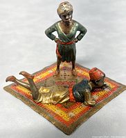 Figurine showing two figures, one standing with hands on hips, the other lying on stomach on a decorated carpet base, front angle.