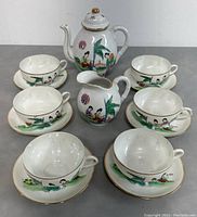 View showing entire tea set: teapot with lid, creamer, and five teacups with saucers, all arranged on a gray surface. Each piece is hand-painted with Chinese female figures, palm leaves, and traditional motifs. Gold accents visible on rims and handles.