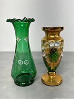 Front view of two Bohemian crystal bud vases side by side, showing green vase with white flower accents and ruffled rim and gold vase with floral lattice decoration.
