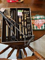 Black carrying case opened showing stainless steel BBQ tools with black handles including spatula, tongs, fork, and skewers. Two green and white checkered pot holders with Bar-B-Que logo are beside the case on a wooden table.