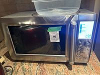 Front view of the Breville microwave showing the stainless steel body, door with dark glass window, label with number sticker, and slightly cracked handle on the right side.