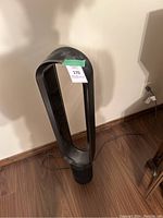 Photo of a black Dyson bladeless tower air conditioner/fan with power cord, placed on a wood floor against a wall. Shows the entire unit and some dust on the surface.