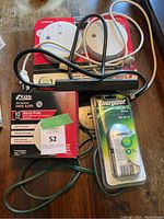 Photo showing two Kidde smoke alarms in packaging, Energizer AA battery pack, black battery charger, multiple extension cords, and power bar laying on wooden floor.
