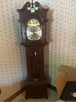 Front view of a wooden grandmother clock showing brass-colored face with Roman numerals, wood case and glass door.