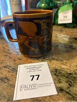 Photo of the brown ceramic mug with Welsh Dragon design on a countertop alongside auction lot number card 77.
