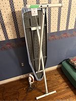 Iron and ironing board standing upright against a wall with diced wallpaper and wooden floor.