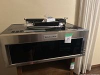 Front view of KitchenAid microwave oven with stainless steel and black glass finish, showing brand label and control panel.