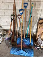 Photo showing the front view of various garden and snow tools lined up against a cinder block wall, including shovels, rakes and blue plastic push snow shovels with wooden handles.