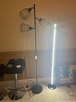 Full view of the black standing lamp with three cage light shades and round base, displayed upright next to a white vertical lamp and a black stool.