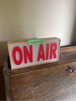 Rectangular light sign with ‘ON AIR’ in large red letters on white panel framed by wood, shown resting on wooden furniture.