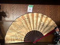 Photo of an oriental folding hand fan with traditional artwork and calligraphy, showing detailed painted landscape and figures in black and brown ink on beige background. Dark ribs and handle are visible.