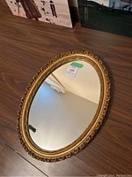 Vintage oval mirror laying flat on a wooden surface, showing detailed gold ornate frame and clear reflective glass.
