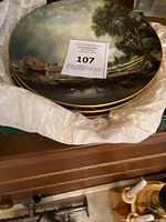 Photo showing folded stack of four decorative plates with a landscape scene, gold trim visible, tissue paper padding underneath.