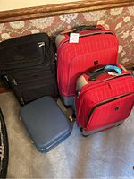 Four suitcases placed on carpeted floor against wallpapered wall: two red hard-shell, one black soft, one blue hard case.