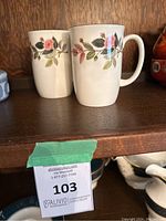 Two white ceramic cups with floral rose pattern and gold trim on a wooden shelf.