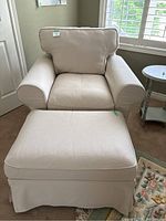 Frontal view of beige slipcovered accent chair behind the ottoman.