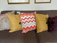 Five accent pillows on a brown couch: multicolor paisley, two mustard yellow with black buttons, and red-white chevron.
