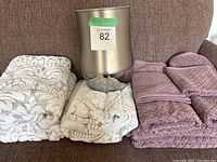 Three towels folded on a couch with a stainless steel garbage bin in the center. Two neutral towels with floral and leaf patterns in a light beige/cream color and one purple quilted towel.