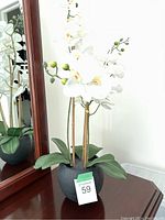 Faux white orchid plant with green leaves in a black pot, shown near a mirror reflecting the plant.