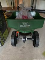 Front view of Scott's green Turf Builder fertilizer spreader showing hopper with fertilizer bag inside and wheels beneath