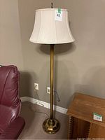 Full view of brass floor lamp with white fabric bell-shaped shade next to a leather chair and wooden table.
