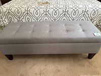 Full front view of the gray upholstered bench showing button tufting on seat and wooden legs, sitting on carpeted floor with a patterned bedspread in the background.
