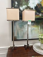 Pair of tall lamps with square beige fabric shades on a wooden surface in front of a modern abstract painting.