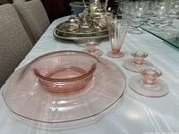 Pink depression glass serving bowl with scalloped edges seen from an angle with three candle holders and one ice cream cup around it on a table.